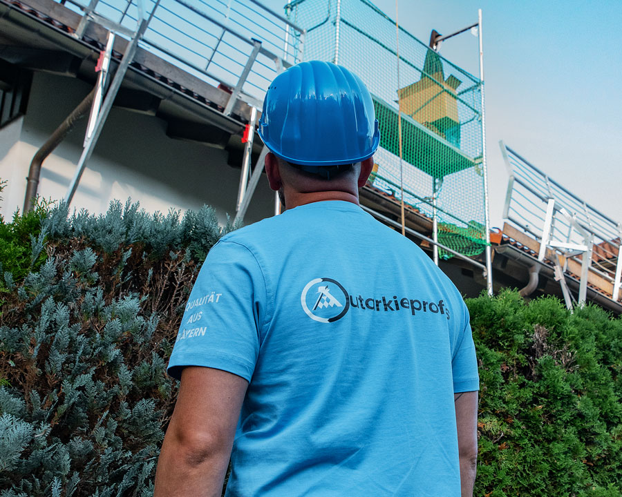 Autarkieprofis-Mitarbeiter mit blauem Schutzhelm blickt auf ein Baugerüst am Haus – Vorbereitung auf Dacharbeiten