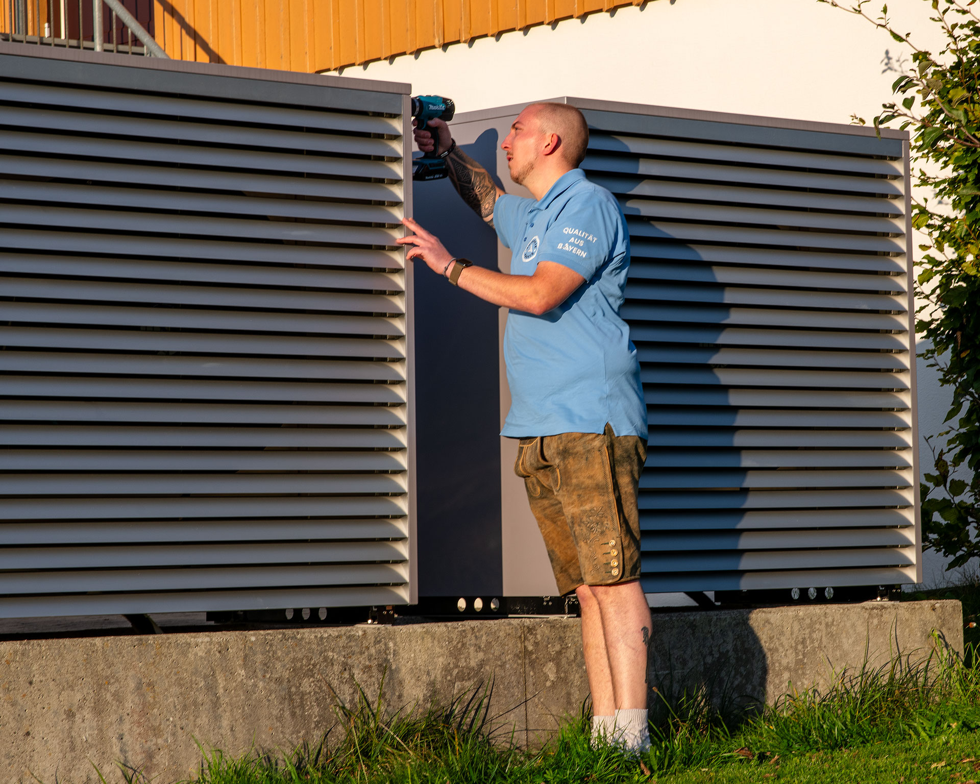 Autarkieprofis-Monteur montiert Aussengeraet einer Waermepumpe mit Akkuschrauber am Haus – Photovoltaik & Heiztechnik, Qualitaet aus Bayern